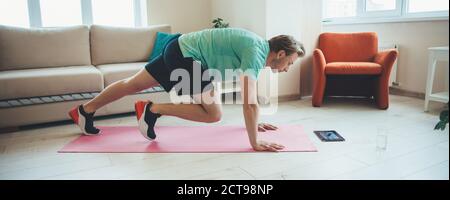Hombre caucásico haciendo ejercicios de fitness en casa usando una tableta mientras planeo sobre una alfombra