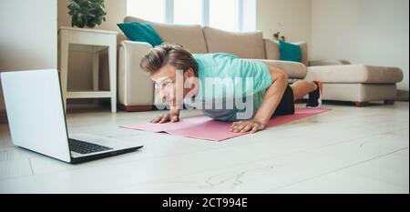 Hombre caucásico haciendo flexiones en casa en el suelo mientras usando un portátil haciendo un gimnasio en casa