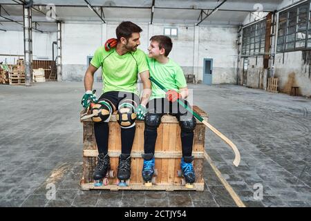 Niño sonriente con brazo alrededor del padre mientras se sentaba con hockey palos en la caja de madera en la cancha