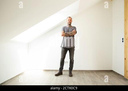 Hombre maduro con los brazos cruzados de pie por la ventana en el ático de nuevo hogar Foto de stock