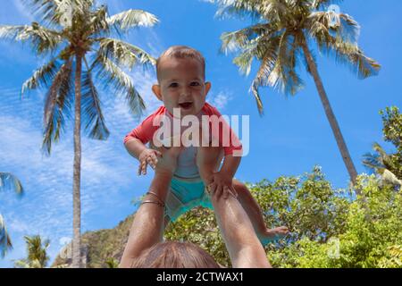 Muy impresionado niño se levantó en los brazos contra el cielo y palmeras Bebé vestido con traje de baño de coral y blanco Fotografía de stock - Alamy