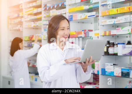 Mujer farmacéutica Asiática usando computadora portátil y lista de verificación inventario o leer información medicamentos en farmacia o farmacia. Medicina, pharmaceut Foto de stock