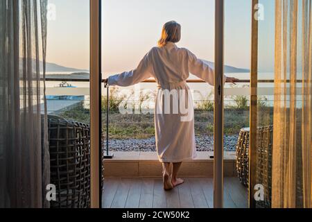Hermosa mujer de vuelta en el albornoz en la terraza del hotel Habitación con vistas al mar Foto de stock