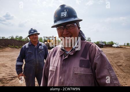 Trabajador de aceite asiático adulto en azul y púrpura ropa de trabajo y azul casco logo BJ (Baker Hughes) posando, sonriendo en la arena del desierto. Depósito de petróleo de Zhayk-Munai