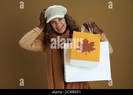 Hola otoño. Sonriente mujer de mediana edad en suéter con guantes de cuero y bolsa de compras de papel sobre fondo de bronce.