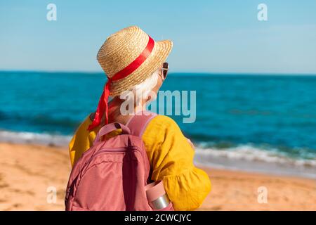 Retrato de una joven feliz en un pajizo de verano sombrero y gafas con mochila turística y termo disfrute de la hermosa vista al mar