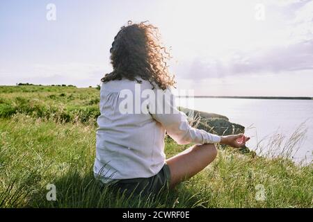 Vista posterior de la mujer de cabello rizado sentada con las piernas cruzadas hierba y meditación en silencio por un lago tranquilo