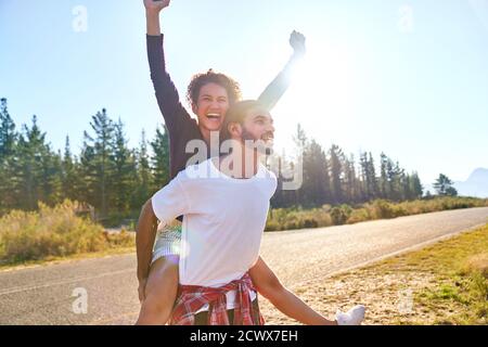 Retrato despreocupado joven pareja piggybacking en la soleada carretera remota