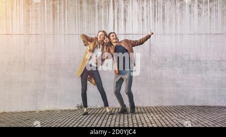 Chica alegre y joven feliz con pelo largo están bailando activamente en una calle al lado de una pared de concreto urbano. Ellos tienen chaqueta de cuero marrón y..
