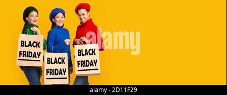 Tres niñas sosteniendo bolsas de compra de viernes negro, fondo amarillo, panorama Foto de stock