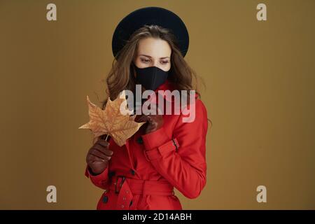 Hola otoño. Mujer de mediana edad moderna y soñadora en abrigo rojo con máscara negra y hoja de arce amarilla otoñal de bronce.
