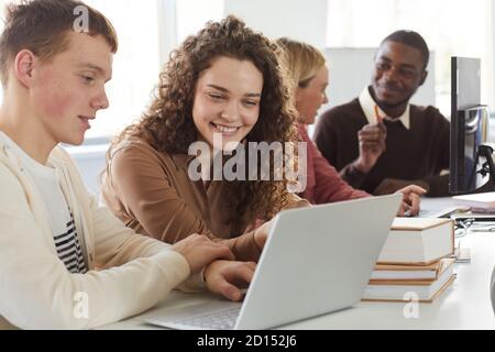 Retrato de una joven sonriente mirando la pantalla del portátil mientras estudiar con un grupo de estudiantes en la biblioteca de la universidad