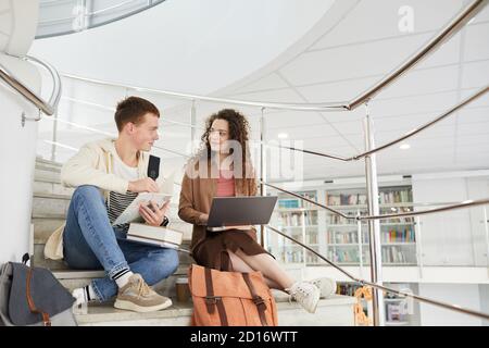 Amplio ángulo de visión en dos estudiantes sentados en las escaleras de la biblioteca de la universidad y usando el portátil mientras trabajan en la tarea, espacio de copia