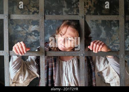 Una joven encarcelada mira tristemente detrás de las rejas en una prisión medieval. Retrato de un criminal en ropa vintage en una fiesta de Halloween
