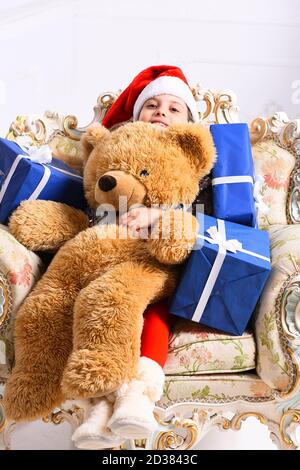 Miss Santa en sombrero rojo se sienta en el sillón chic y tiene cajas de regalo y un oso de juguete enorme. El adorable niño recibe regalos. Navidad y concepto sorpresa. Chica con cara feliz sobre fondo blanco.