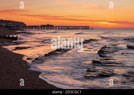 El sol se levanta detrás del muelle de Hastings en el este de Sussex con el Marea retrocediendo en la playa de Saint Leonards revelando las rocas Foto de stock
