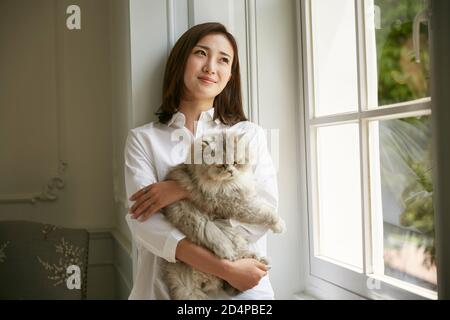 hermosa joven asiática de pie junto a la ventana en la casa de celebración un gato en los brazos mirando serena y contento