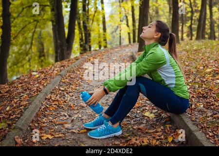 Corredor descansando después de entrenar en el parque de otoño. Mujer cansada sosteniendo una botella de agua. Estilo de vida activo deportivo
