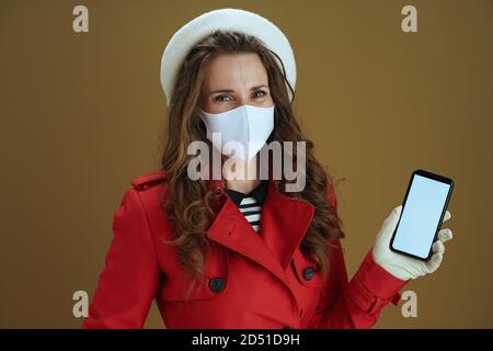 Hola otoño. Retrato de la feliz mujer de mediana edad moderna en un abrigo rojo mostrando pantalla en blanco smartphone contra fondo beige.