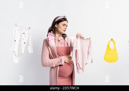mujer triste y embarazada colgando ropa de bebé en la línea de ropa en blanco Foto de stock