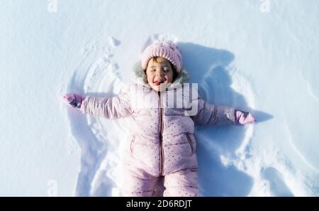 Vista superior retrato de una niña pequeña y alegre tumbada en la nieve en la naturaleza invernal, haciendo ángeles. Foto de stock