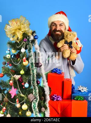 Guy en red hat con cajas de regalo de navidad y el oso de peluche en las manos. Viernes negro antes de Navidad el concepto. El hombre con la barba y el rostro alegre sobre fondo azul. Santa con rojo presenta cerca de abeto decorado.