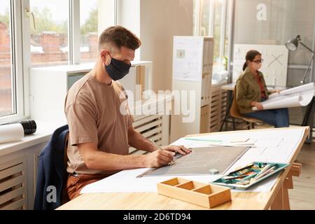 Vista lateral retrato de arquitecto barbudo maduro con máscara mientras se sienta en el escritorio de dibujo en la luz del sol, espacio de copia