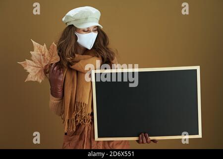 Hola otoño. Mujer moderna de mediana edad en suéter con máscara médica blanca y hojas de arce amarillo otoño mostrando pizarra blanca aislada en beis ba