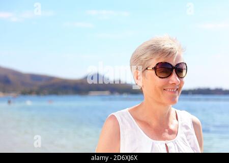 Mujer mayor sonriente en la playa. Mujer anciana feliz en los casuales está usando gafas de sol. La mujer está en sus vacaciones de verano