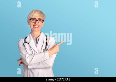 Mujer sonriente doctor en gafas, abrigo blanco con estetoscopio apunta el dedo al espacio libre para un buen tratamiento Foto de stock