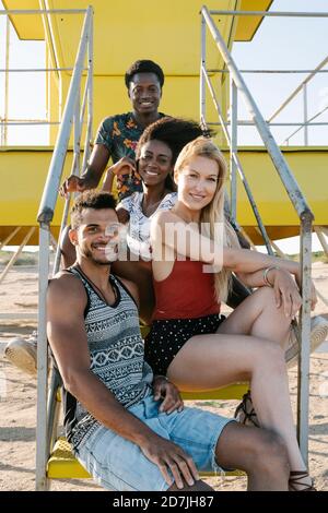 Amigos jóvenes sonriendo sentados a pasos de socorrista choza playa durante el día soleado Foto de stock