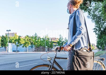 Joven caucásico vestido con una cara azul máscara sosteniendo una bolsa para portátil y sosteniendo un estilo retro bicicleta mirando a la carretera antes de cros