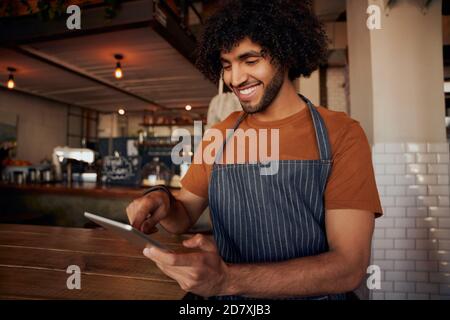 Feliz joven camarero con delantal sosteniendo y usando digital tableta en el café