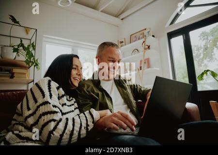 Hermosa pareja joven y cariñosa mirando el portátil y sonriendo mientras sentados juntos en el sofá Foto de stock