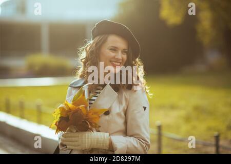 Hola otoño. Sonriente mujer de mediana edad en abrigo de trinchera beige y bereta negra con hojas amarillas de otoño fuera en la ciudad en otoño.