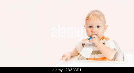 Un niño pequeño y feliz de ojos azules está sentado en la mesa y tomando agua de una botella con un chupete. Independiente. Espacio de copia Foto de stock
