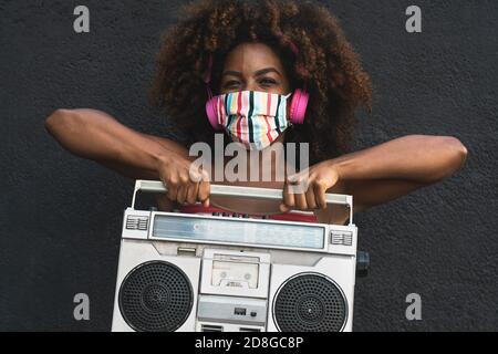 Joven mujer afro con máscara bailando al aire libre mientras escucha a la música con auriculares inalámbricos y boombox clásico estéreo Foto de stock