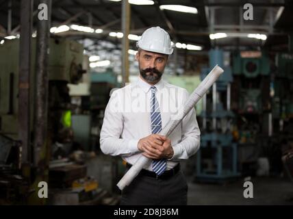 El director de hipster con barba con sombrero de ingeniero de pie contra la máquina en producción en fábrica Foto de stock