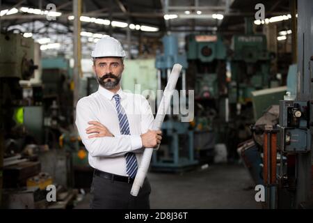 El director de hipster con barba con sombrero de ingeniero de pie contra la máquina en producción en fábrica Foto de stock