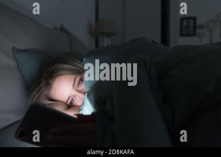 Mujer cansada y soñolienta acostada en la cama bajo la manta usando un teléfono inteligente a última hora de la noche, no puede dormir. Insomnio, nomofobia, concepto de trastorno del sueño. Social Foto de stock