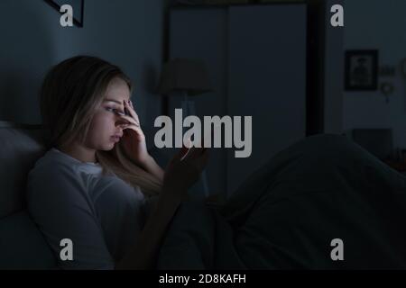 Una joven mujer cansada y soñolienta acostada en la cama usando un smartphone y tocando su frente, no puede dormir. Insomnio, nomofobia, concepto de trastorno del sueño. Gad Foto de stock