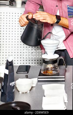 Las manos masculinas del barista preparan el café vertiendo agua hirviendo de una caldera negra en un embudo blanco de un matraz de vidrio. Foto de stock