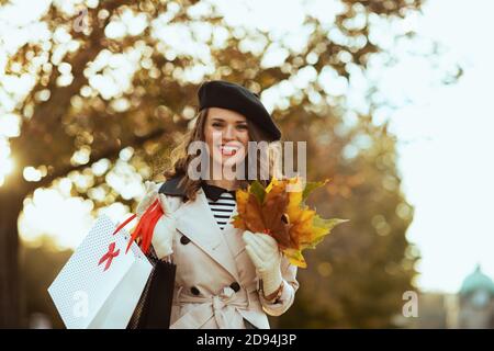 Hola otoño. Sonriente mujer de mediana edad en abrigo de trinchera beige con bolsas de compras y hojas amarillas de otoño al aire libre en la calle de la ciudad en otoño.