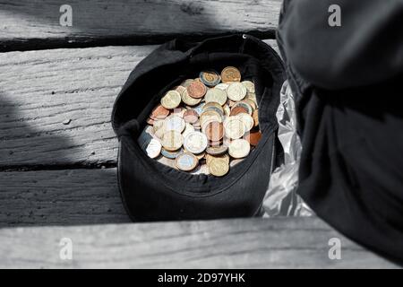 Un mendigo se sienta cerca de una gorra con monedas y pide limosnas. Gorra con unas monedas de limosna Foto de stock