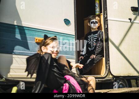 Hermana y hermano llevando disfraces de Halloween y sentados en frente de un autocaravana Foto de stock