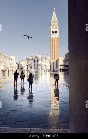 Turistas tomando fotografías en la Piazza San Marco con marea alta, acqua alta. La Basílica de San Marcos y el campanario se reflejan en el agua.