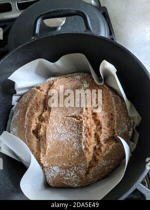 Pan redondo recién hecho en la sartén negra para horno de hierro fundido con una tapa en el lateral. Pan casero con cortes en el lado que se pone en un pergamino pa