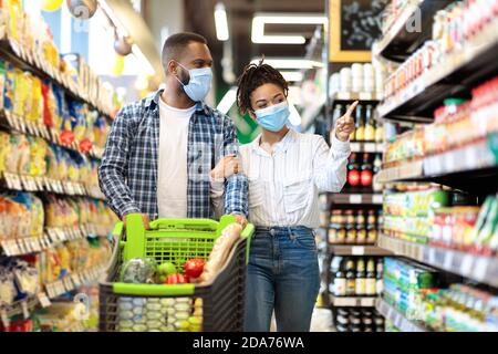 Pareja Africana en máscaras protectoras en supermercado haciendo compras de comestibles Foto de stock
