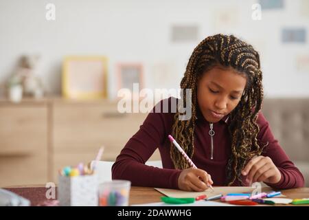 Vista frontal retrato de una adolescente afroamericana haciendo la tarea mientras se sentaba en el escritorio en el interior de la casa, espacio para copias