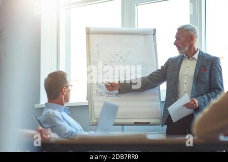 Un hombre de negocios maduro y seguro que apunta al rotafolios y explica algo a sus colegas mientras se celebra una reunión de intercambio de ideas en la oficina. Foto de stock